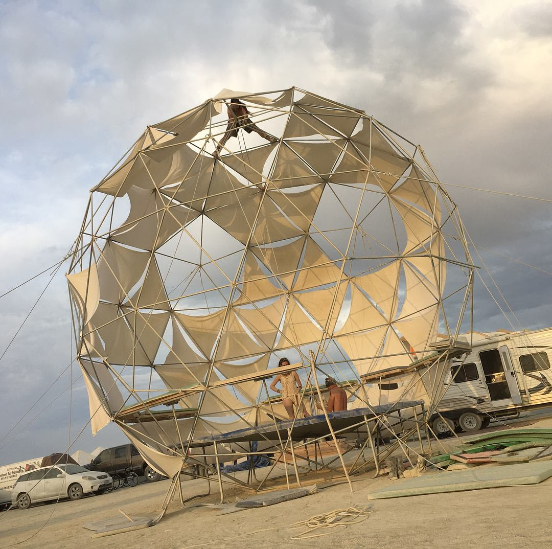 A ~24-foot sphere of metal pipes and cloth triangles with a trampoline installed in the base, with three campmates working on it. p/c Amanda Tay.