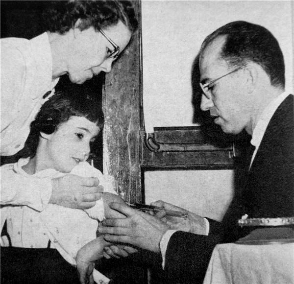 Photograph of Jonas Salk giving a vaccine injection to a school-aged girl, assisted by an adult woman.